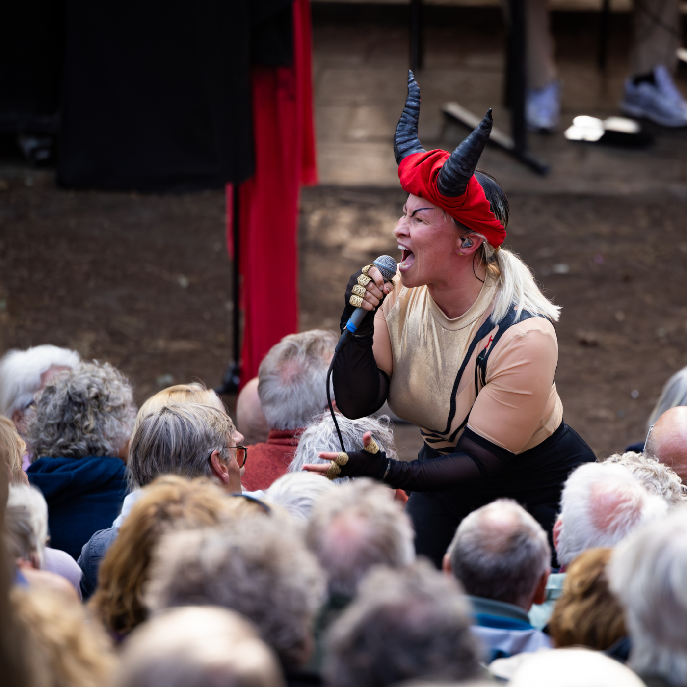 Performer met duivelskostuum en zwarte hoorns zingt in microfoon te midden van zittend publiek tijdens buitenvoorstelling.