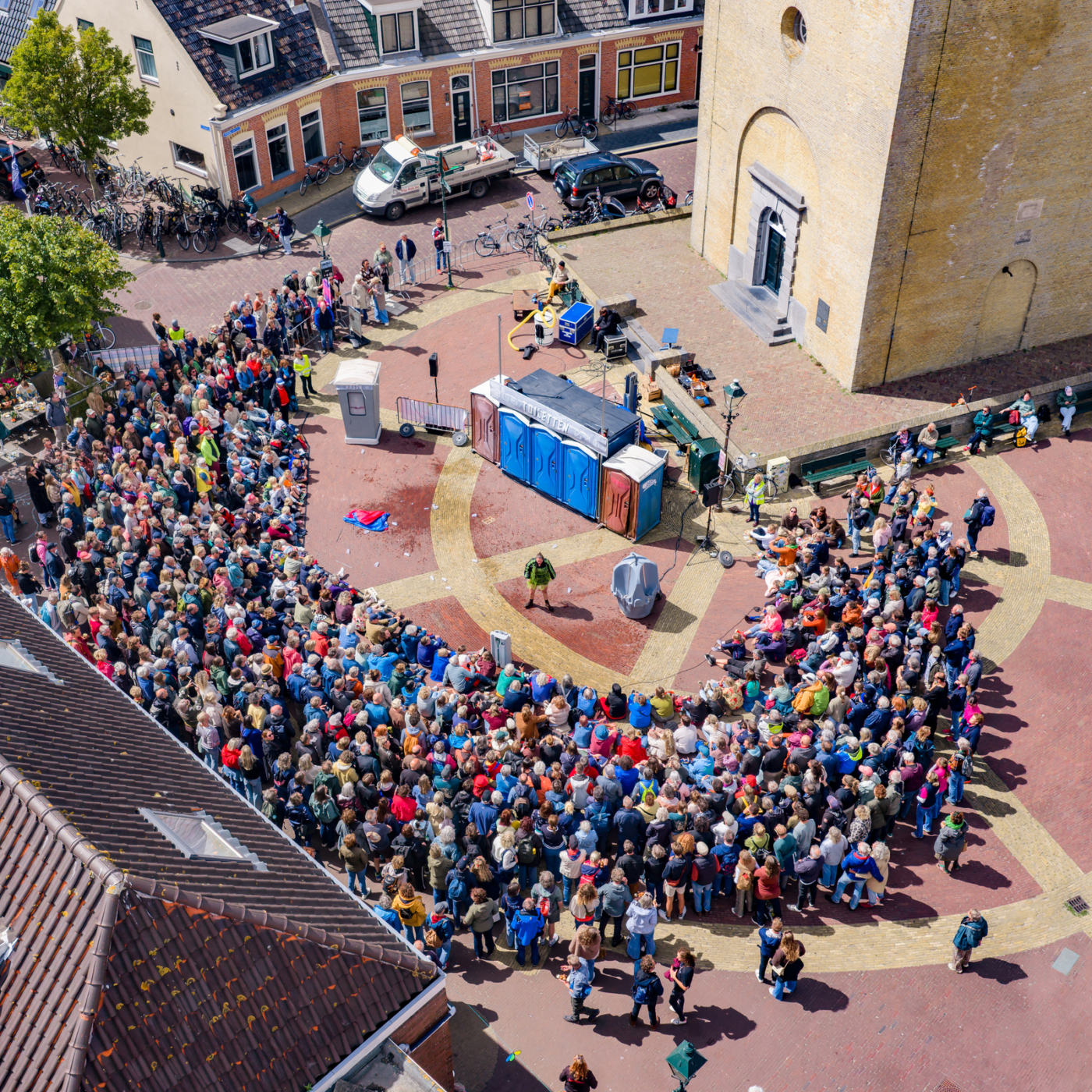 Luchtfoto van grote mensenmassa op een plein bij een kerk tijdens het Oerol Festival op Terschelling, met straatartiesten in het midden.