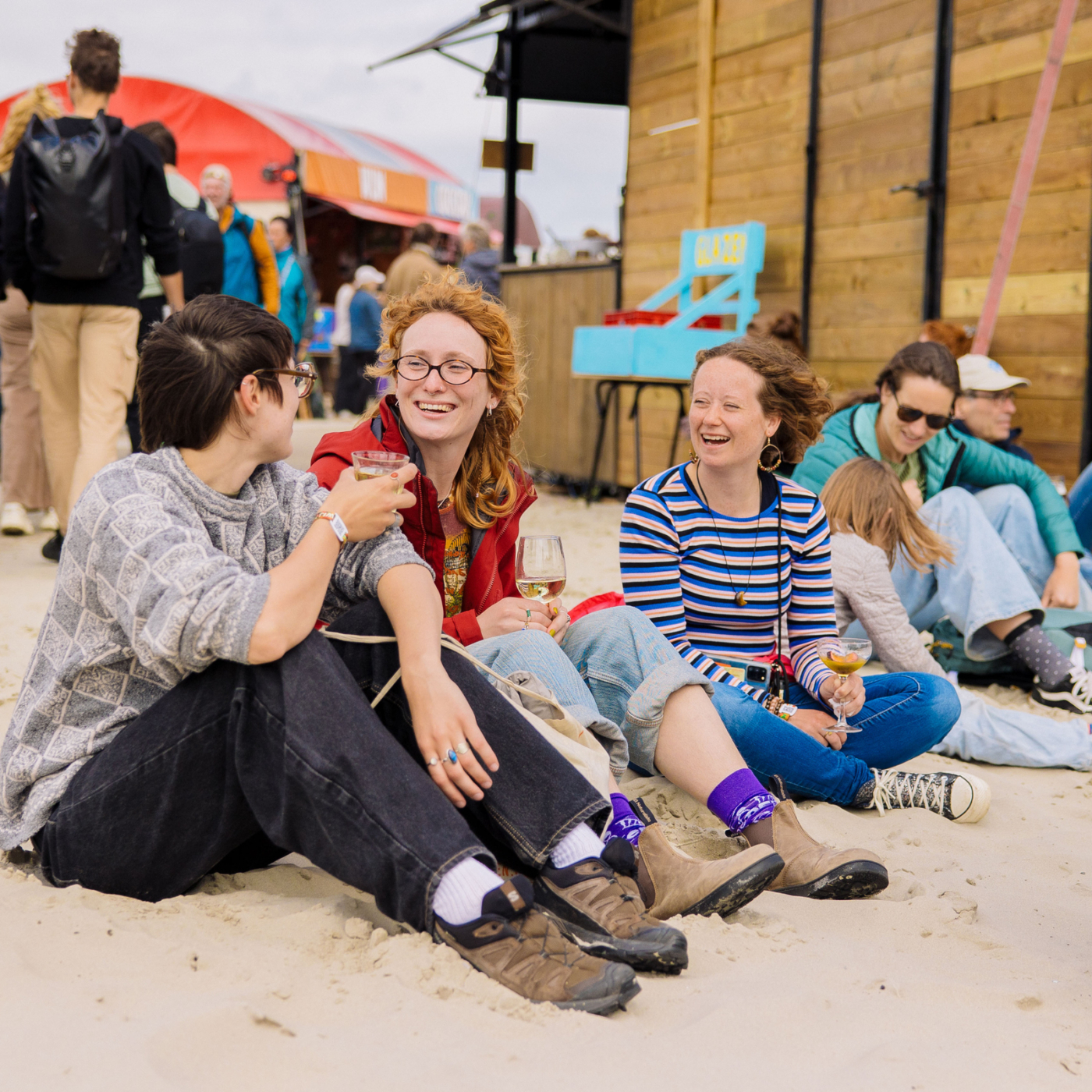 Drie lachende vrouwen zitten op het zand tijdens Oerol Festival op Terschelling, met wijnglazen in hand en festival op de achtergrond.