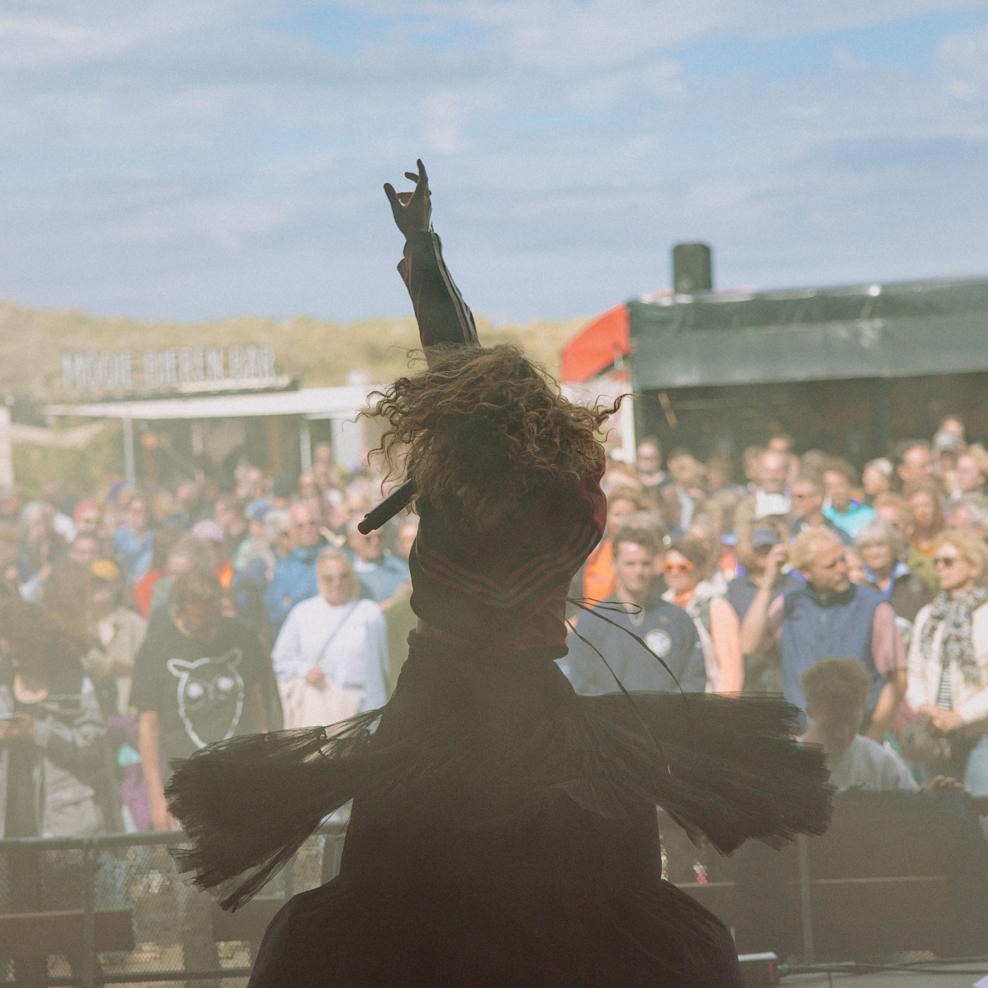 Artiest met krullend haar en zwarte outfit steekt arm omhoog op buitenpodium voor grote festivalmenigte onder blauwe lucht.