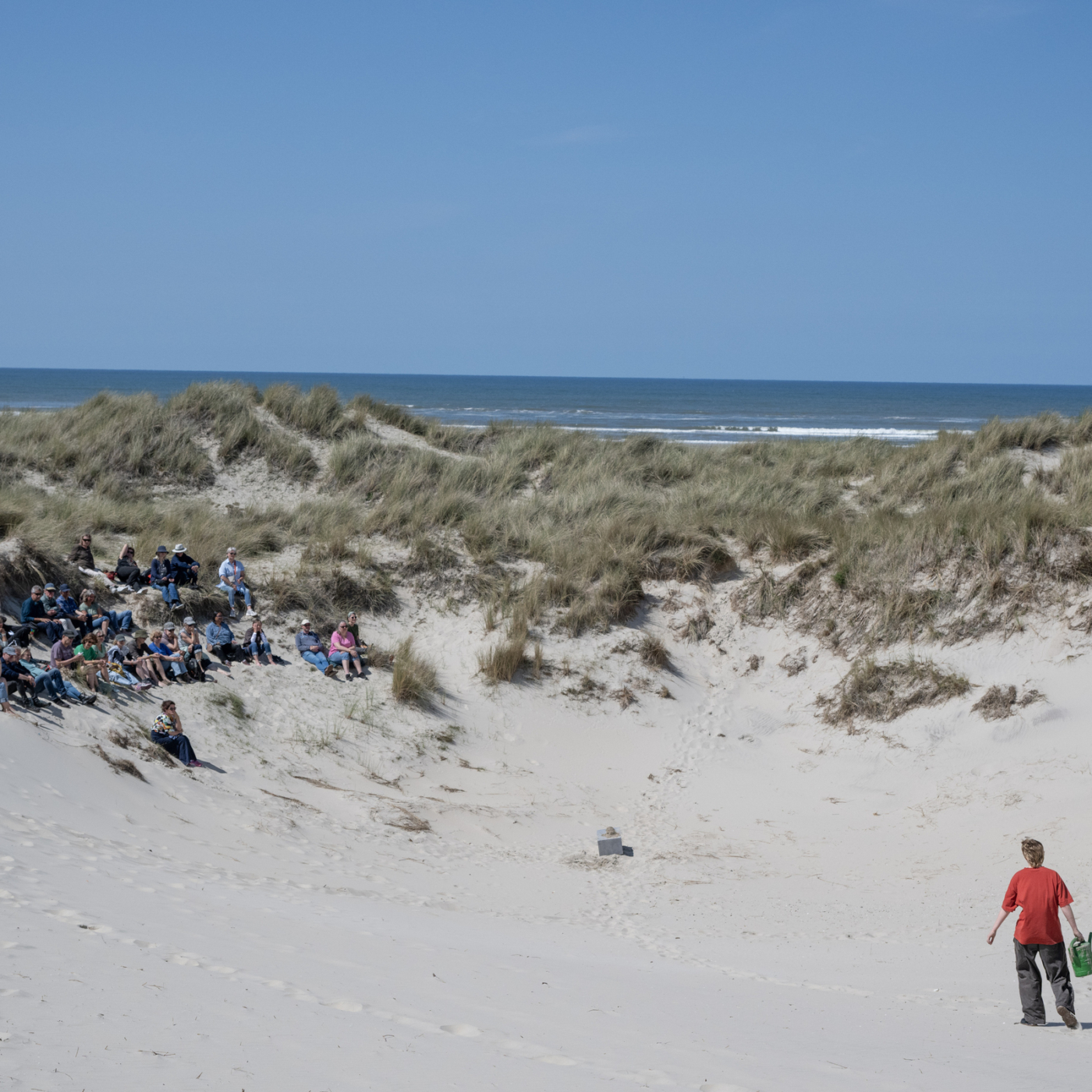 Groep mensen zit op begroeide duinhellingen langs de Noordzeekust, terwijl een kind in rood shirt over het strand loopt.