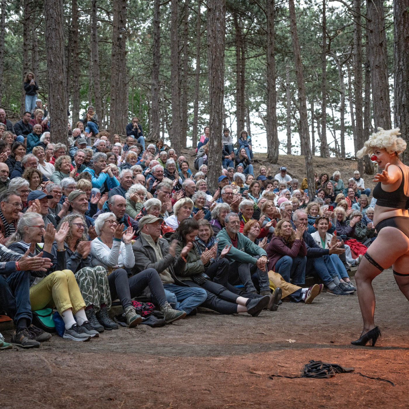 Clown in lingerie en blonde pruik treedt op voor klappend publiek in bosrijke buitenomgeving tijdens openluchtvoorstelling.