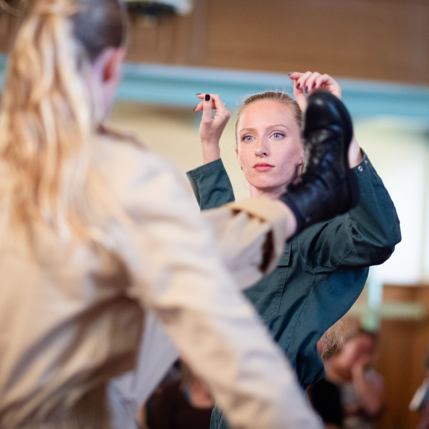 Danseres in groen pak houdt een been hoog boven haar hoofd tijdens een repetitie, terwijl een andere danser op de voorgrond onscherp zichtbaar is.