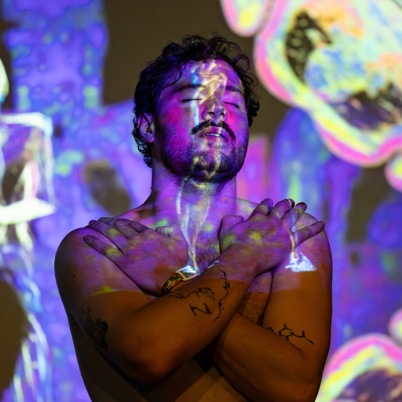 Shirtloze man met gesloten ogen en gekruiste armen, omgeven door kleurrijke psychedelische projecties in paars, roze en geel.