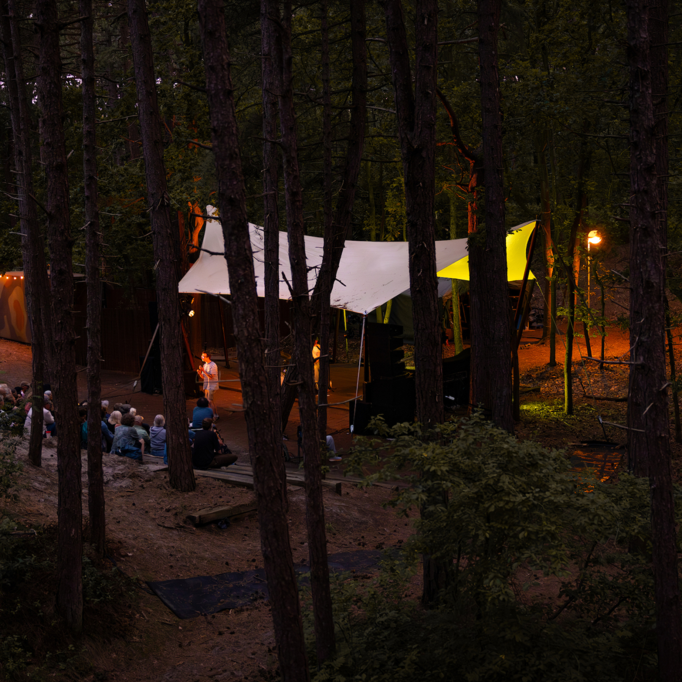 Buitenvoorstelling in donker bos bij avond: publiek zit op de grond voor verlicht podium met witte zeildoeken tussen de bomen.
