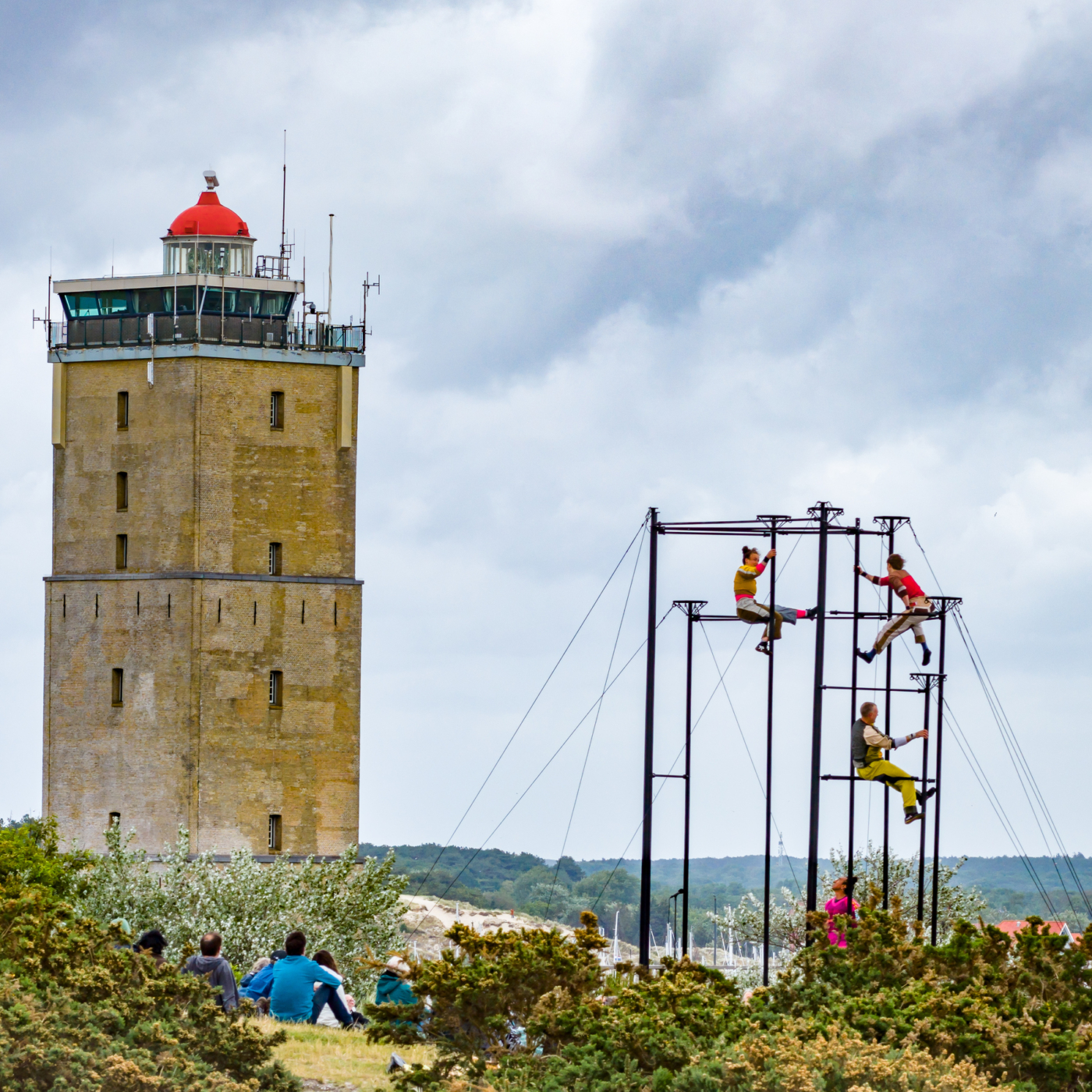 Acrobaten klimmen op een stalen constructie tijdens Oerol Festival, met de vuurtoren van Terschelling op de achtergrond en toeschouwers in de duinen.