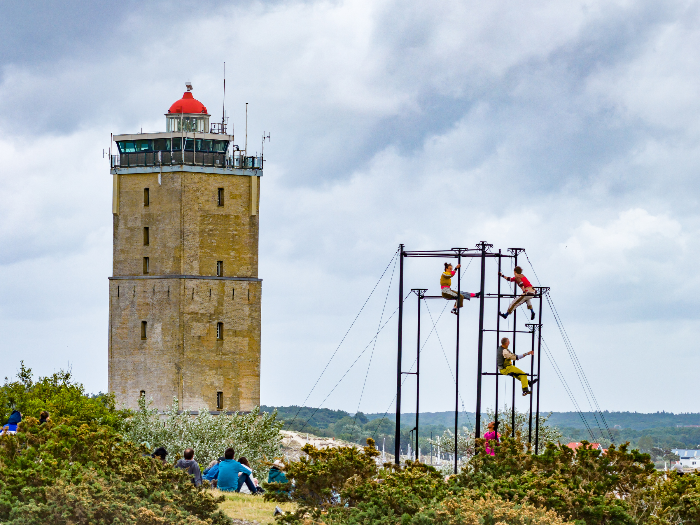 Acrobaten klimmen op een stalen constructie tijdens Oerol Festival, met de vuurtoren van Terschelling op de achtergrond en toeschouwers in de duinen.
