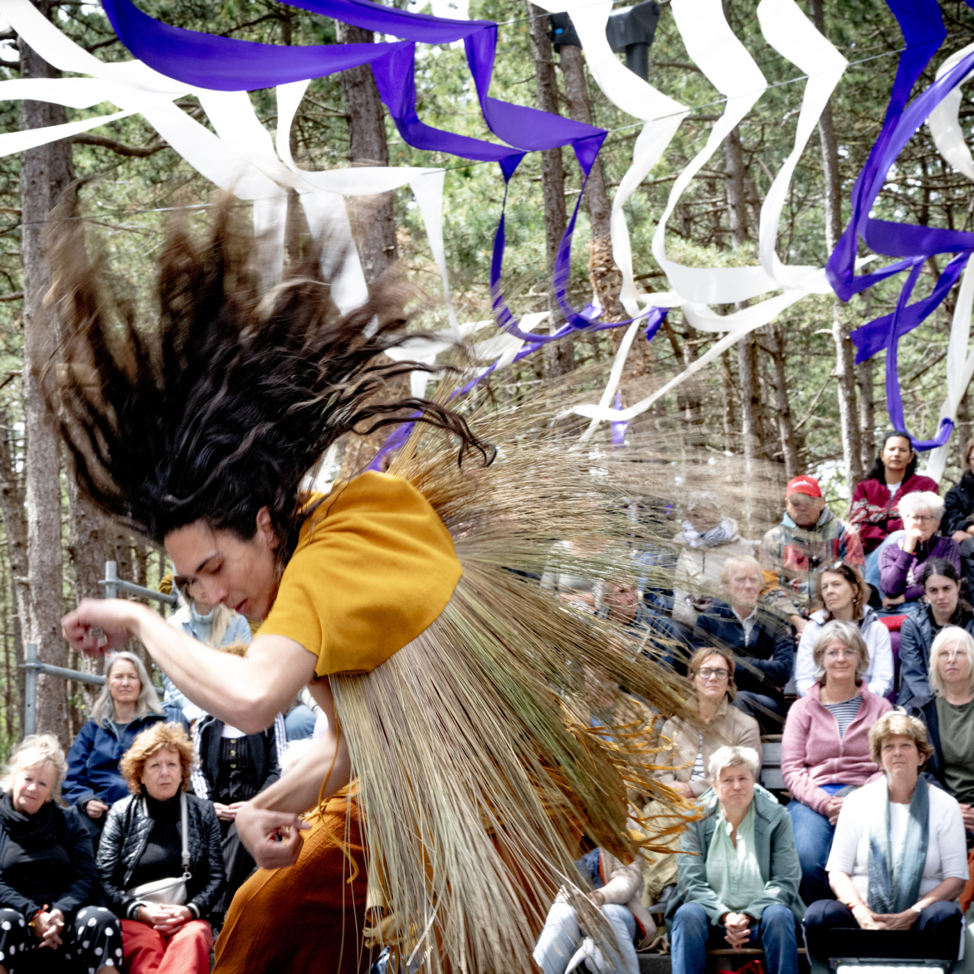 Danser in oker outfit met vliegend haar en strogras beweegt dynamisch voor publiek in bostheater met paarse en witte linten.