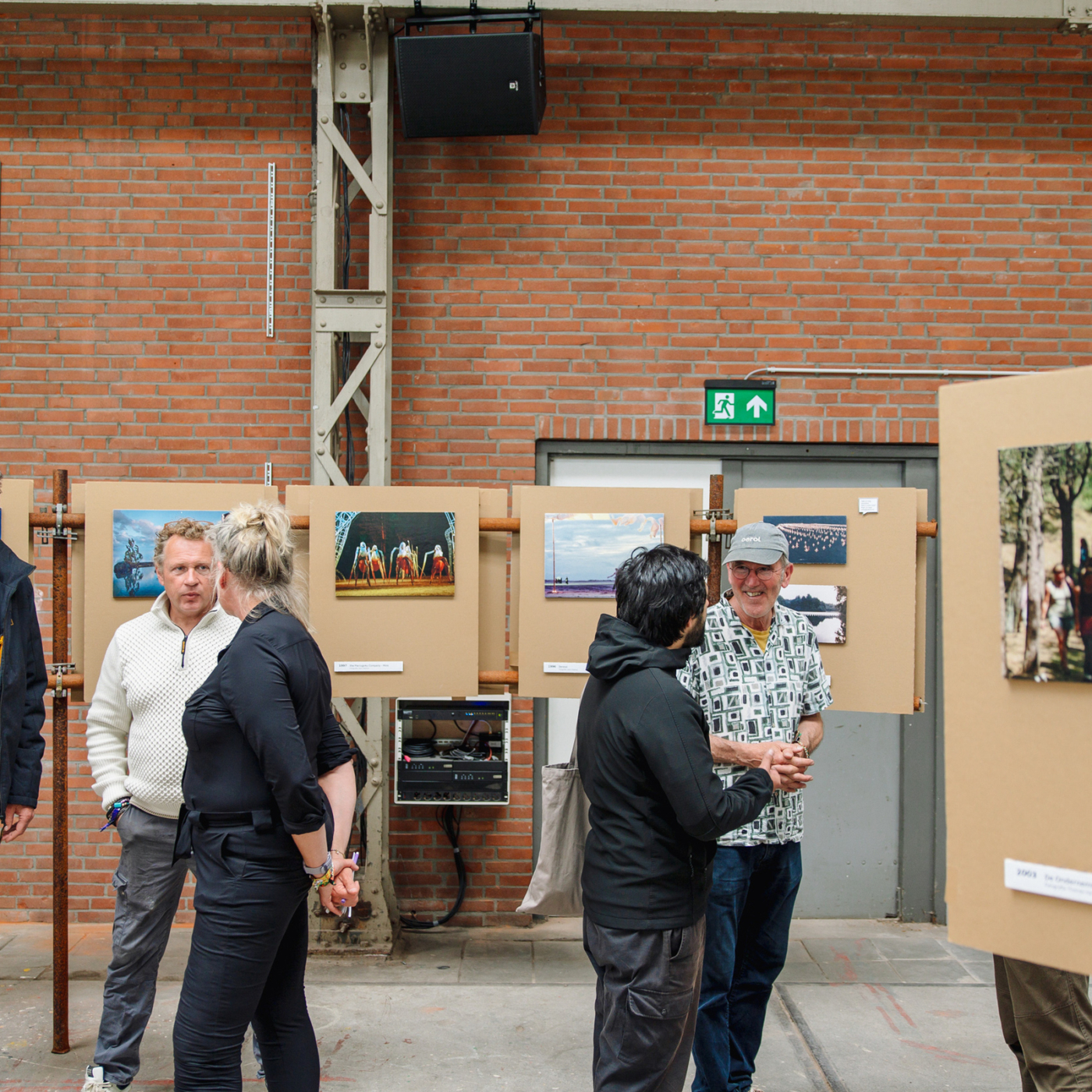 Bezoekers bekijken en bespreken foto's op panelen tijdens een buitententoonstelling tegen een rode bakstenen muur.