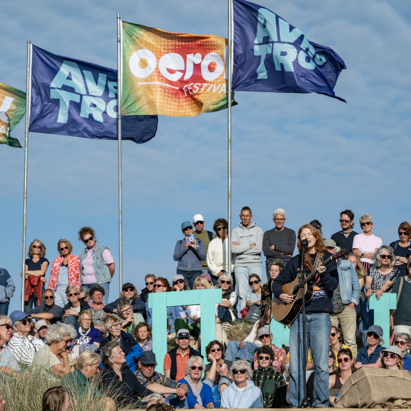 Zangeres speelt gitaar op het Oero Festival met AVROTROS-vlaggen en een grote menigte toeschouwers in de duinen.