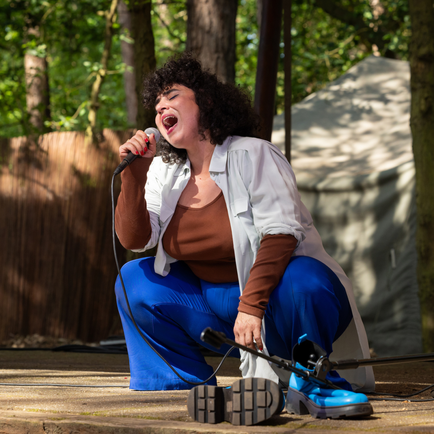 Meral Polat, een zangeres met krullend haar, zingt geknield op buitenpodium, microfoon in hand, blauwe broek en witte blouse, bomen op achtergrond.