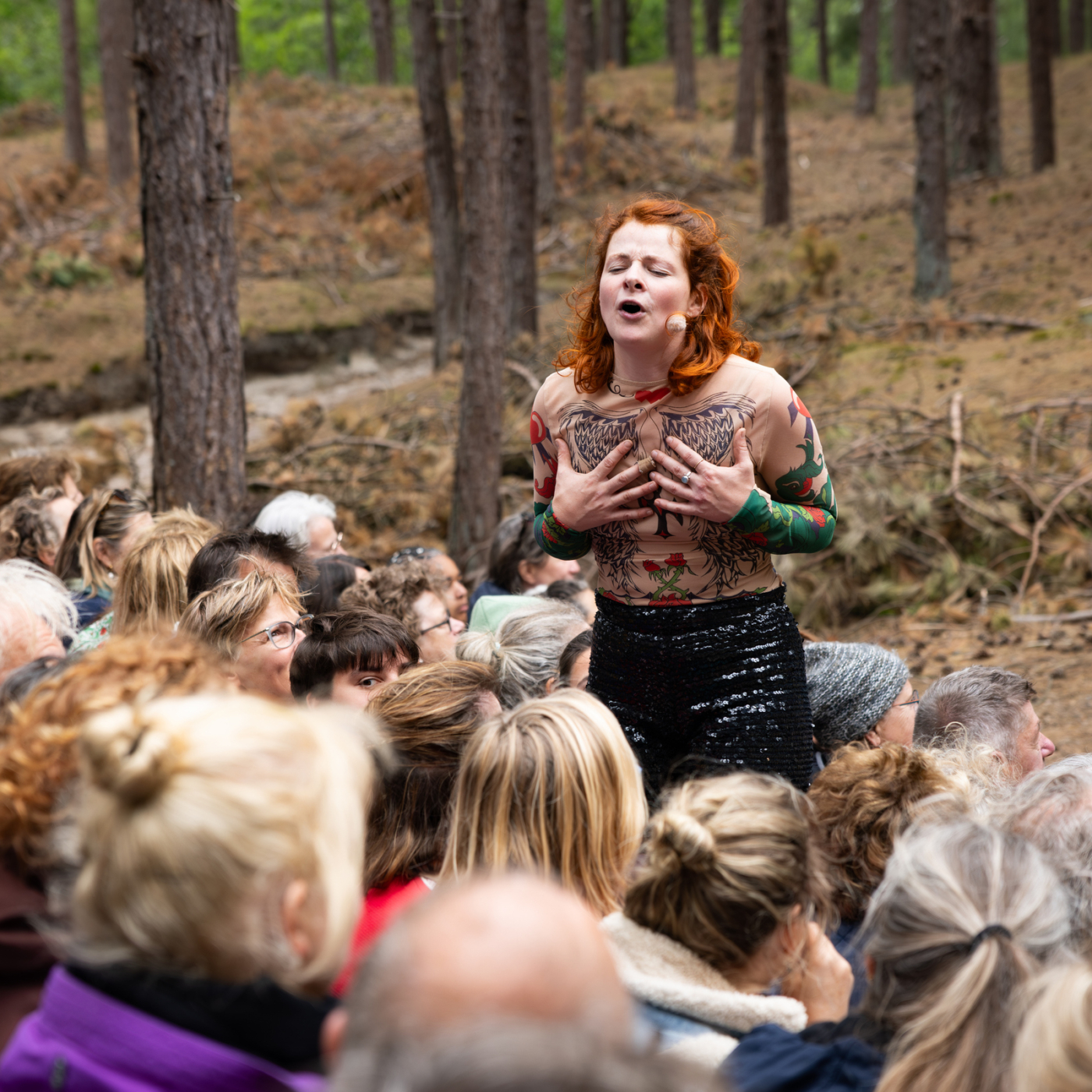 Roodharige performer met shirt aan waardoor het lijkt alsof ze tatoeages heeft, zingt met gesloten ogen temidden van een dicht publiek in een dennenbos.
