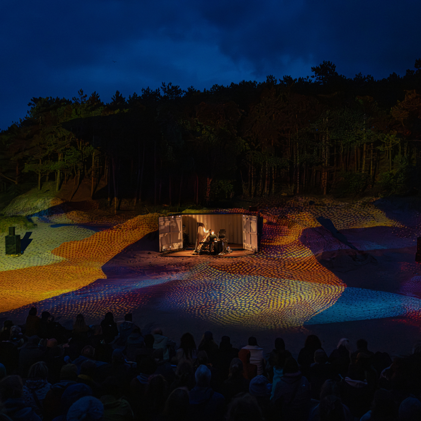 Buitenpodium in nachtelijk boslandschap met kleurrijke lichtprojectie op zandheuvels, publiek kijkt naar muzikant in verlichte container.