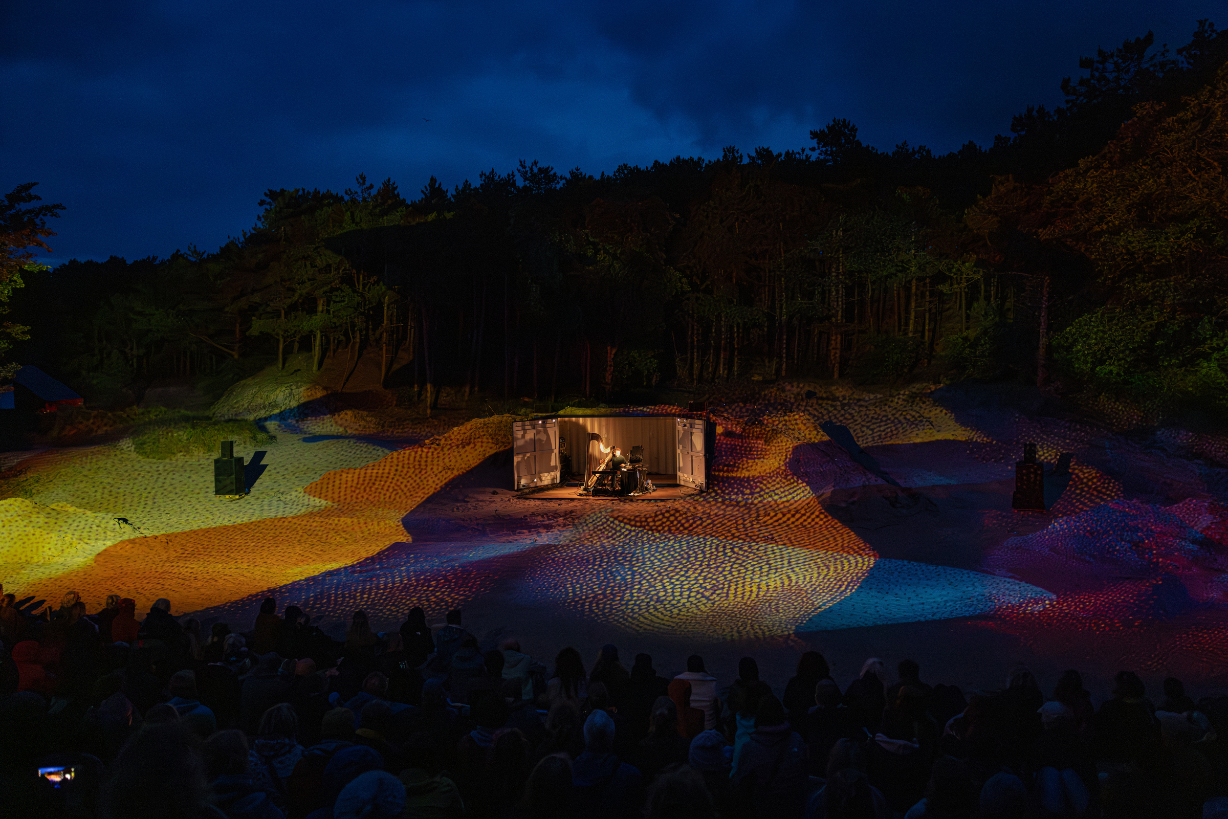 Buitenpodium in nachtelijk boslandschap met kleurrijke lichtprojectie op zandheuvels, publiek kijkt naar muzikant in verlichte container.