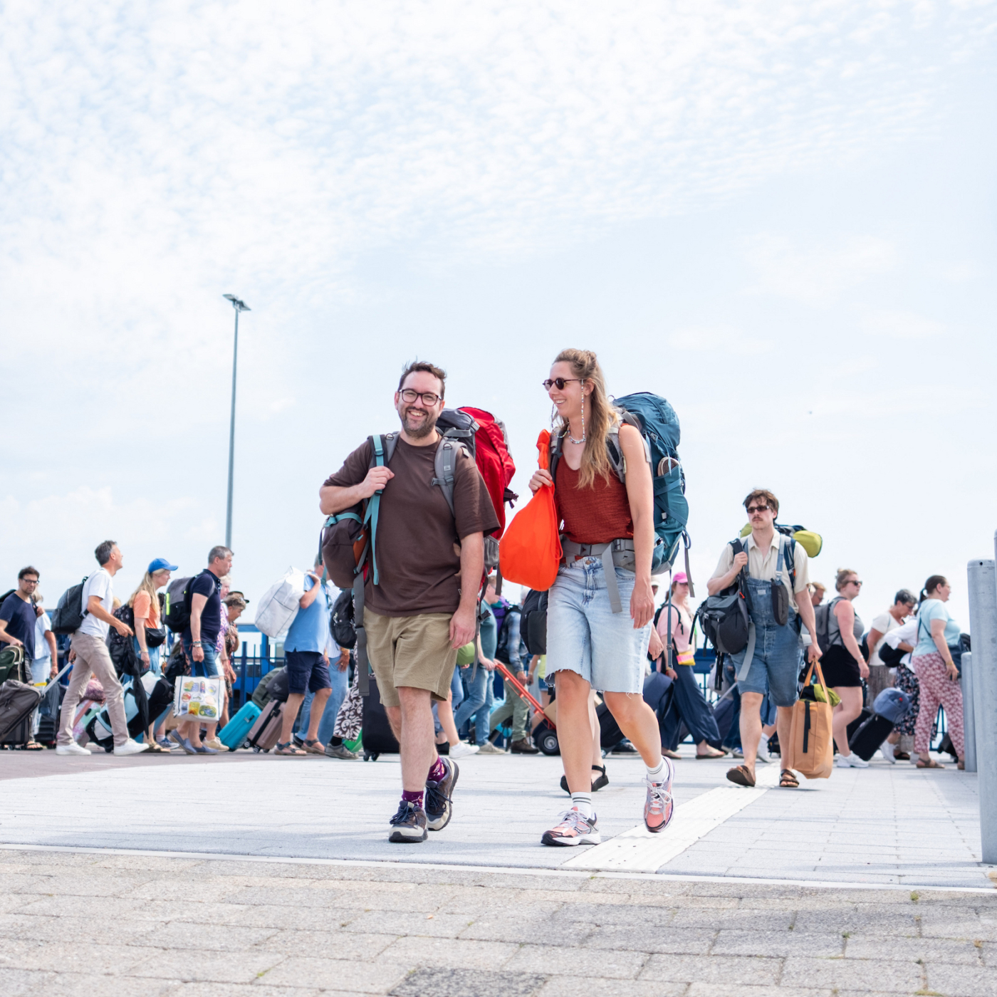 Lachend stel met grote backpacks loopt over een aanlegsteiger, omringd door drukte van meereizende toeristen met koffers.