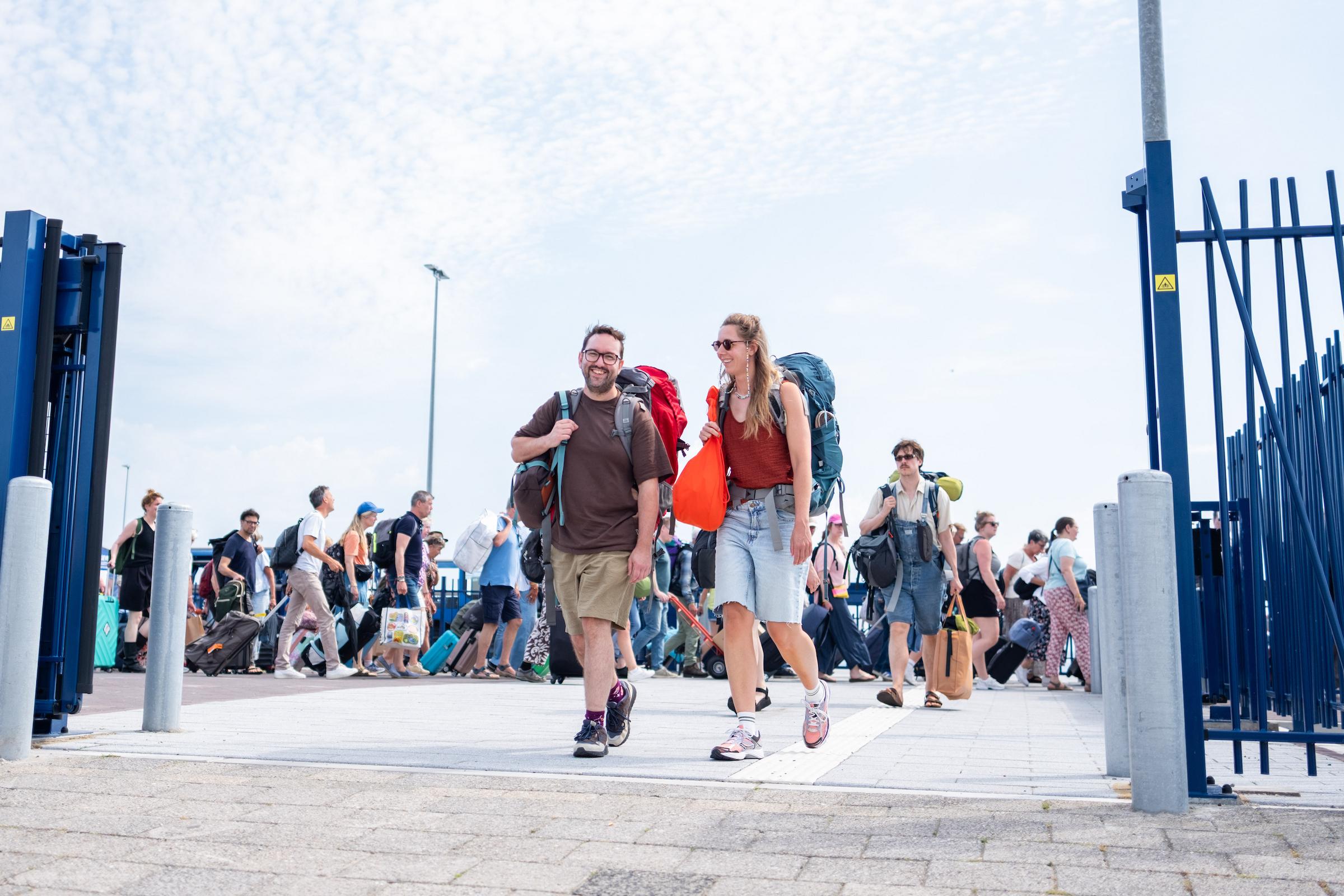Lachend stel met grote backpacks loopt over een aanlegsteiger, omringd door drukte van meereizende toeristen met koffers.