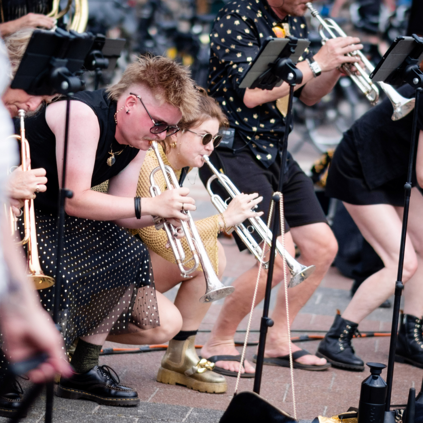 Twee trompettisten hurken tijdens een buitenoptreden, omringd door andere muzikanten met koperblazers en notenstandaards.
