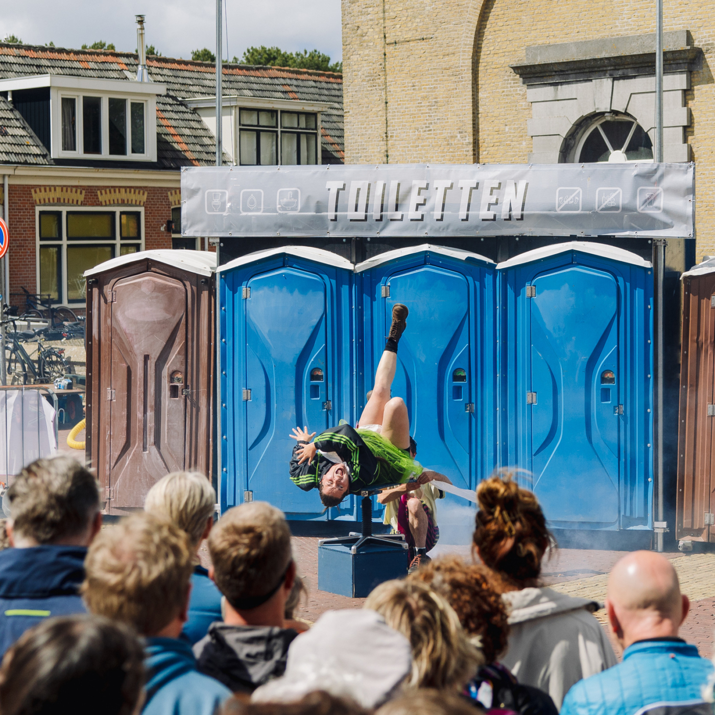 Straatartiest voert acrobatische handstand uit voor publiek, met rij blauwe draagbare toiletten op de achtergrond tijdens buitenevenement.