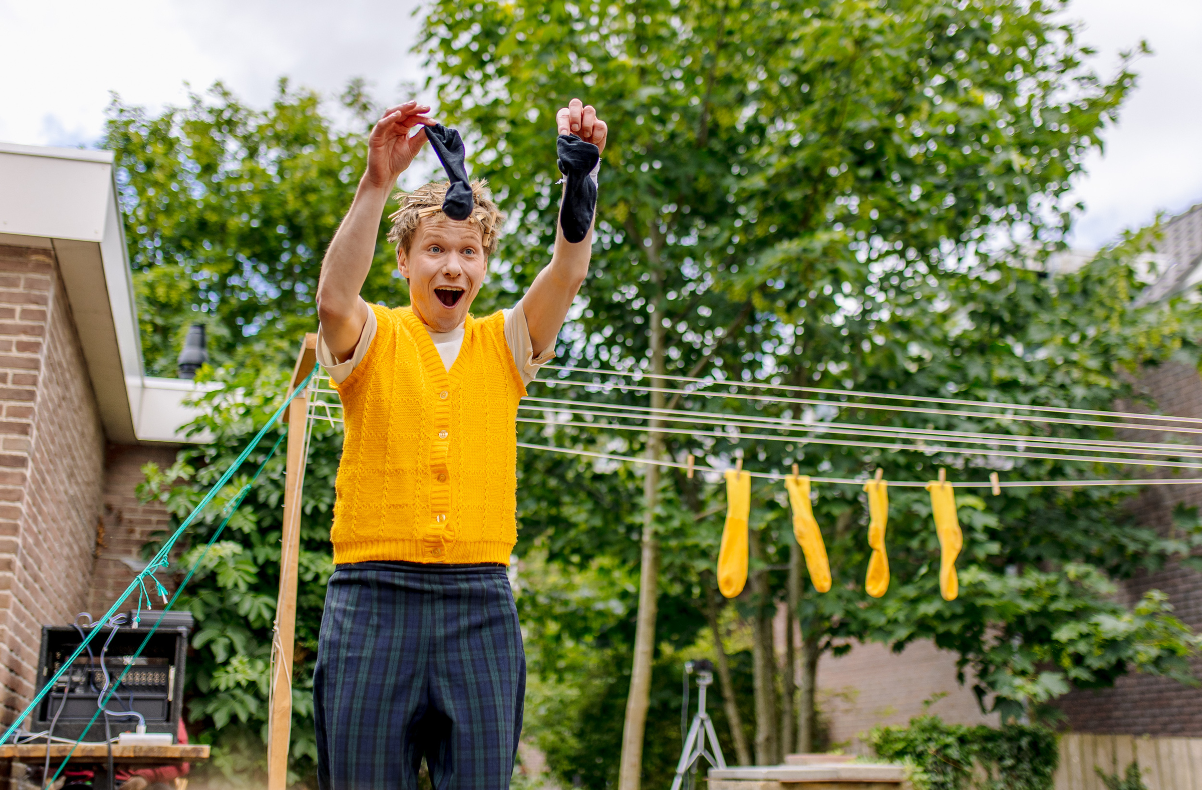 Enthousiaste man in geel vest houdt twee zwarte sokken omhoog, op de achtergrond hangen gele sokken aan een droogrek buiten.