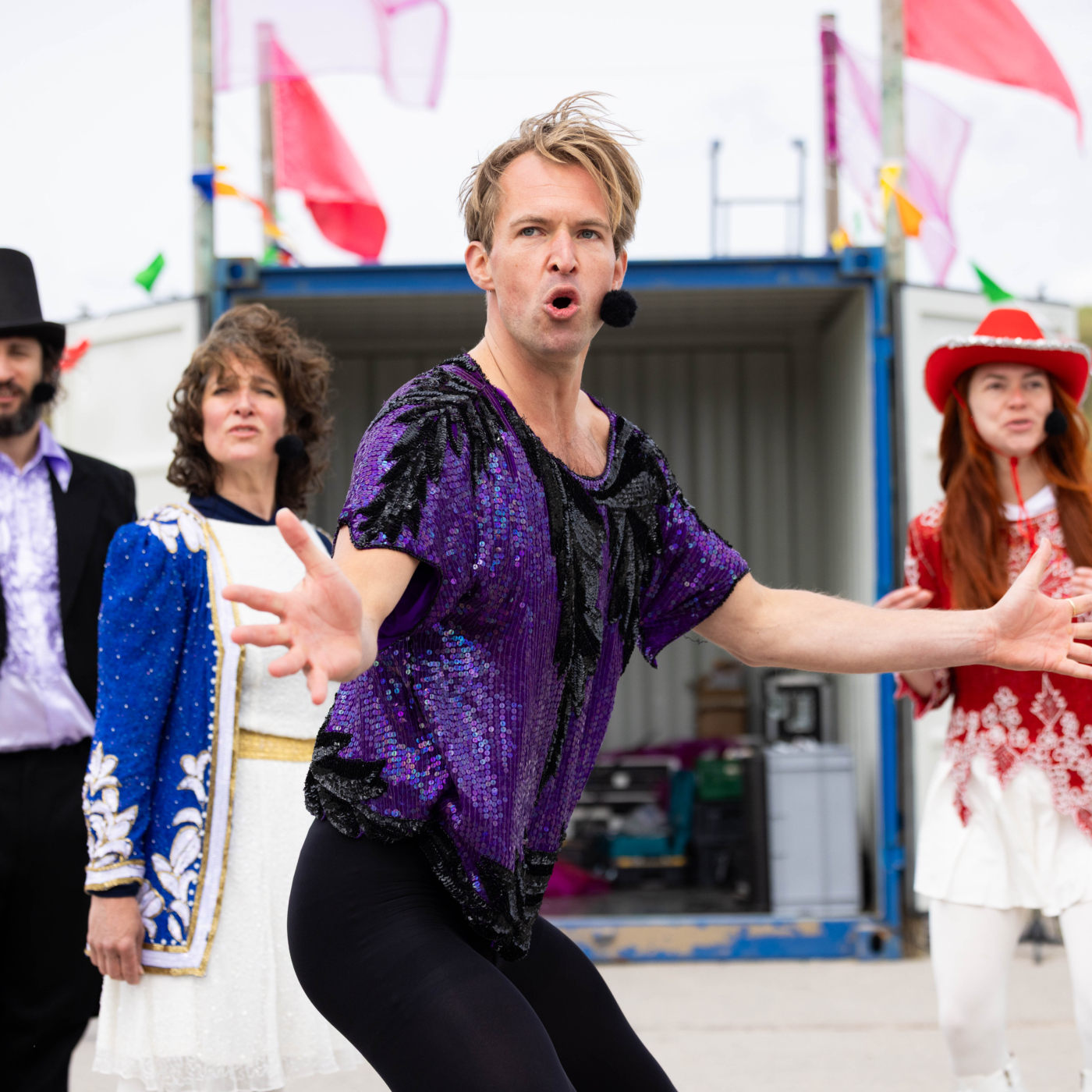Performer in paars paillettenkostuum zingt expressief op buitenpodium, omringd door drie medespelers in kleurrijke theaterkostuums tijdens Oerol.