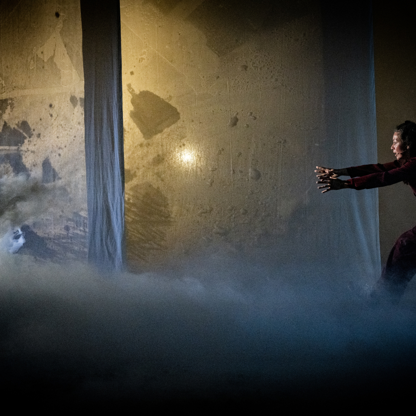 Theaterperformance met danseres in rood kostuum die armen uitstrekt in mistige scène met verlichte doeken op de achtergrond.