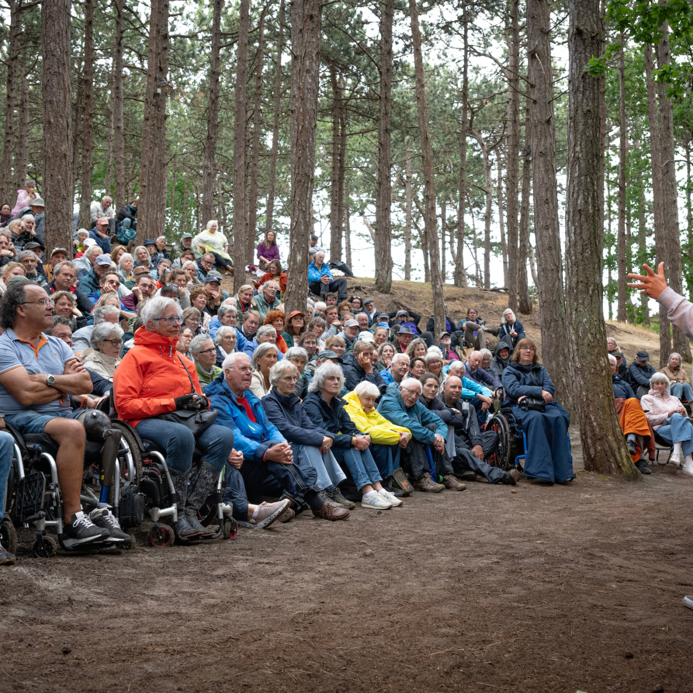 Grote groep bezoekers, waaronder rolstoelgebruikers, luistert naar een spreker in een dennenbos tijdens een buitenevenement.