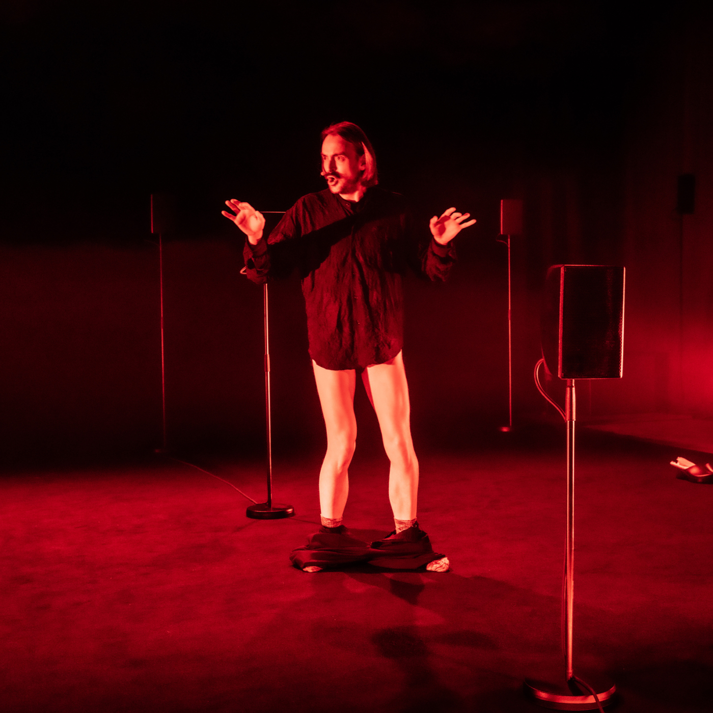 Performer in zwart kostuum gebaart expressief op roodverlicht podium omringd door luidsprekers tijdens Oerol Festival op Terschelling.