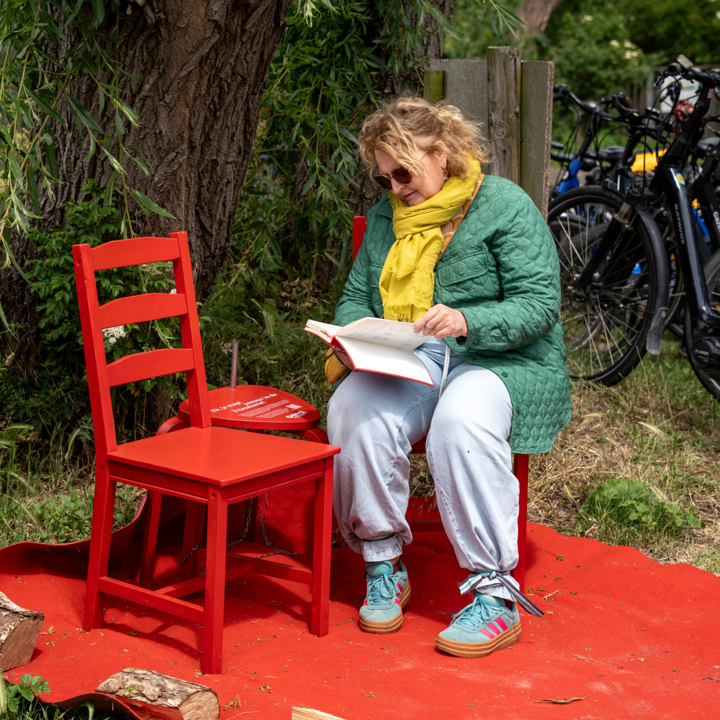 Vrouw met gele sjaal en groene jas leest een boek op rode stoel buiten, omringd door bomen en fietsen.