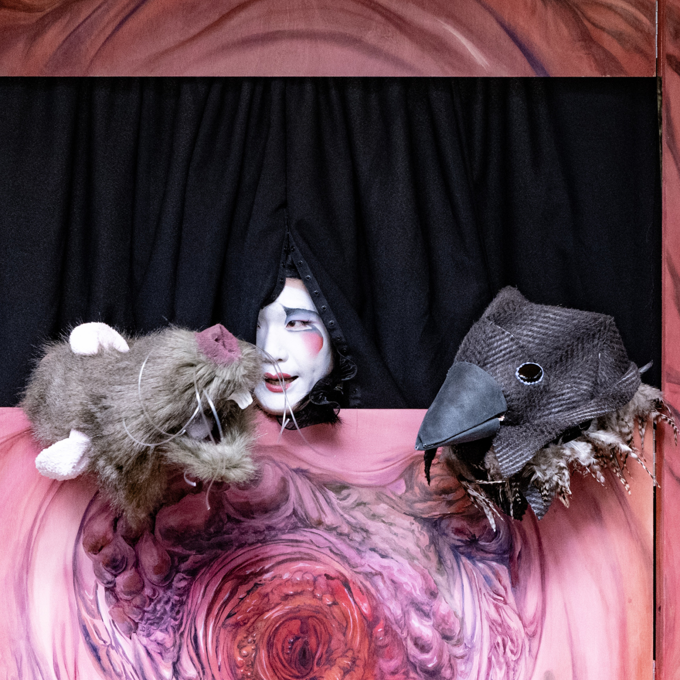 Gemaskerde performer met wit geschminkt gezicht kijkt tussen zwart gordijn door, geflankeerd door rat- en vogelpop op kleurrijk beschilderd podium bij Oerol Festival.