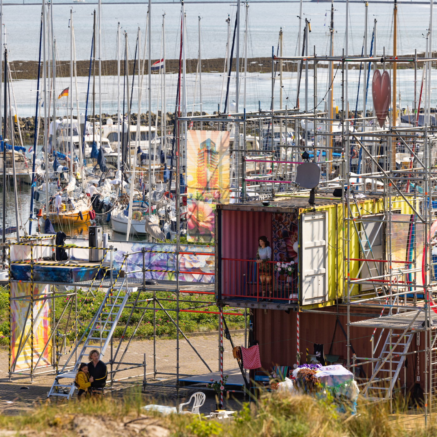 Kleurrijke zeecontainers met steigers en graffiti-kunst in een jachthaven, met mensen op het podium en zeilboten op de achtergrond.