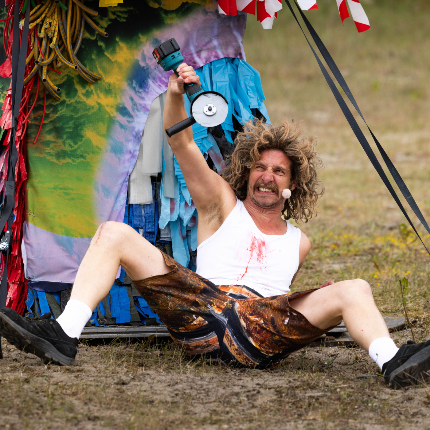 Theatrale performer zit op de grond met slijptol omhoog, draagt wit hemd met rode vlekken en kleurrijke broek, tijdens buitenvoorstelling.